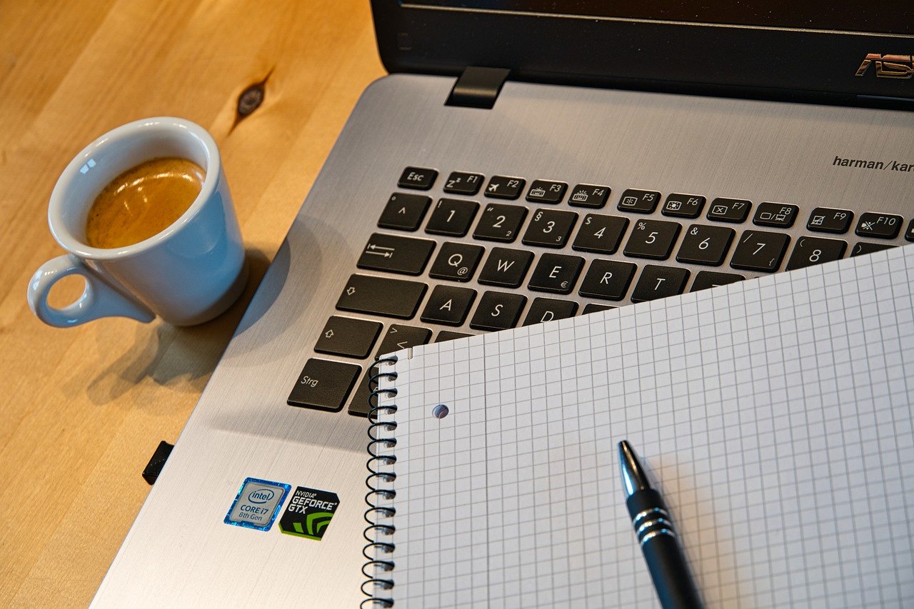 Part of a laptop, a notebook, pen and cup of tea on table background