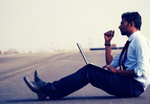 Indian man in white shirt and black trousers and tie sitting on the street with laptop